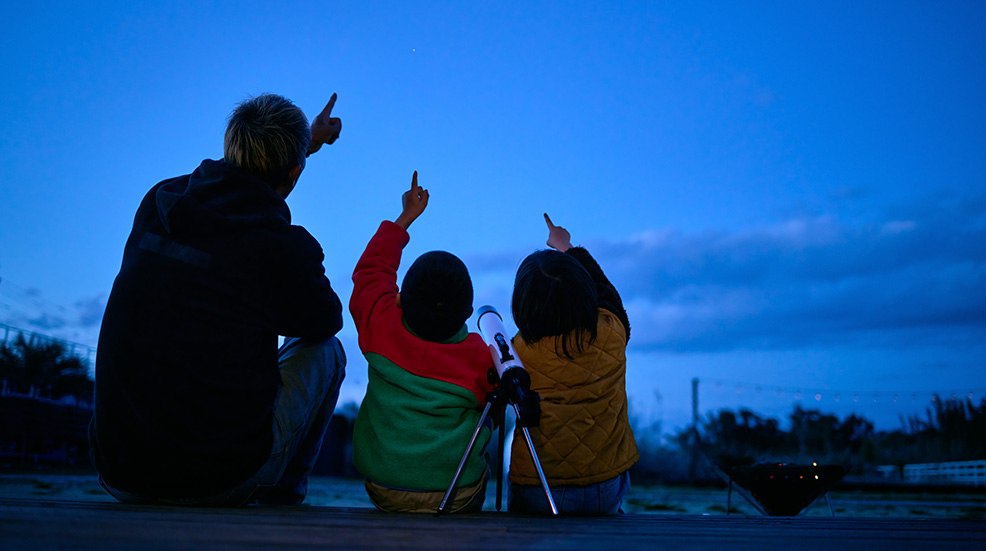 Slhouette of father and two children pointing up at the sky with a telescope between them